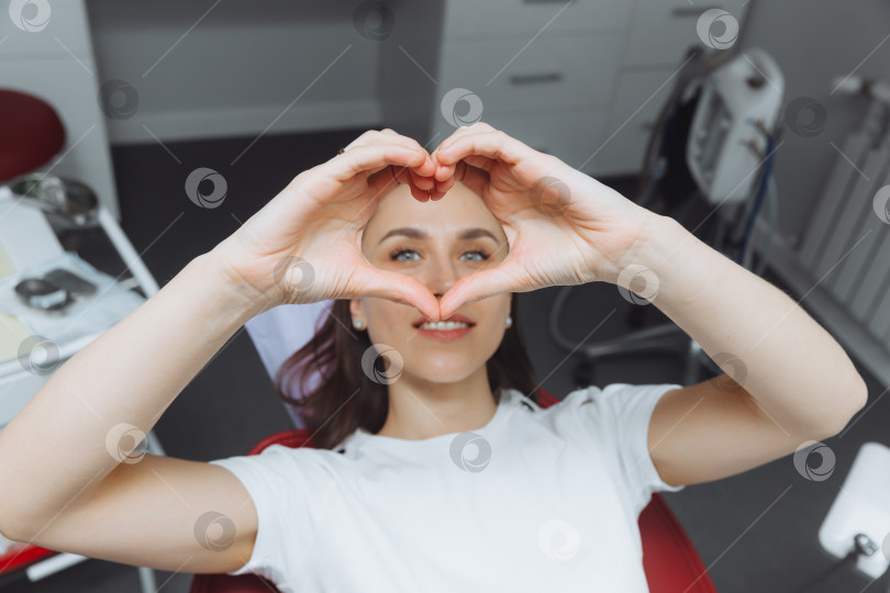 Скачать улыбающаяся молодая женщина, показывающая сердце руками в кресле стоматолога. фотосток Ozero