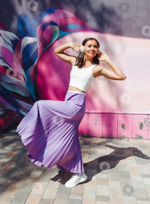 Скачать молодая возбужденная женщина в топе и юбке, в наушниках, слушает музыку, гуляет, танцует с поднятыми руками на открытом воздухе на фоне розовой стены. фотосток Ozero