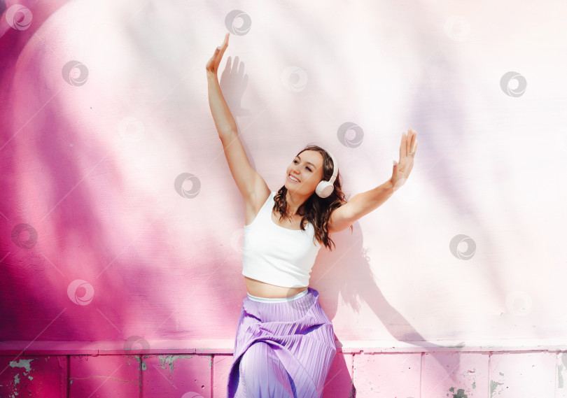 Скачать Молодая счастливая кавказка, одетая в белый топ и фиолетовую юбку, в наушниках, слушает музыку, танцует на городской улице у стены здания. Концепция образа жизни городской молодежи. фотосток Ozero