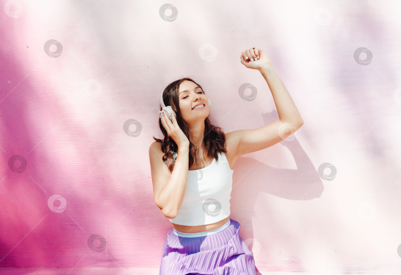 Скачать Молодая счастливая кавказка, одетая в белый топ и фиолетовую юбку, в наушниках, слушает музыку, танцует на городской улице у стены здания. Концепция образа жизни городской молодежи. фотосток Ozero