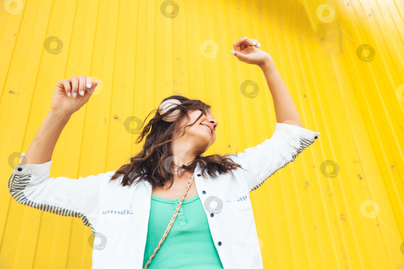 Скачать Изображение красивой жизнерадостной девушки в полный рост, прыгающей во время прослушивания мелодии, выделяющейся на ярко-желтом фоне. молодая женщина в наушниках танцует на улице. поколение z фотосток Ozero