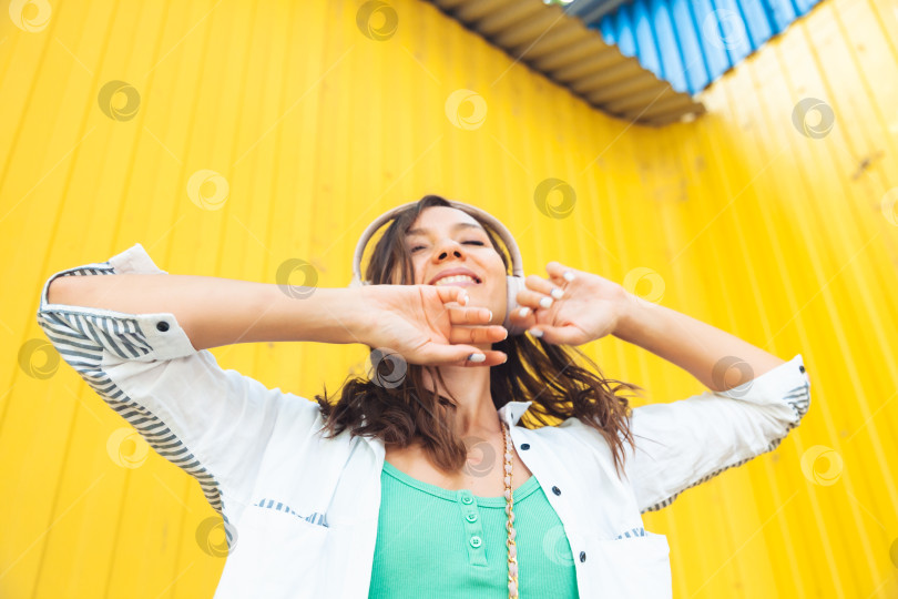 Скачать Изображение красивой жизнерадостной девушки в полный рост, прыгающей во время прослушивания мелодии, выделяющейся на ярко-желтом фоне. молодая женщина в наушниках танцует на улице. поколение z фотосток Ozero
