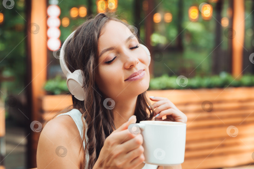 Скачать Молодая красивая женщина в наушниках сидит за столиком в летнем кафе и пьет кофе или чай. поколение z. фотосток Ozero