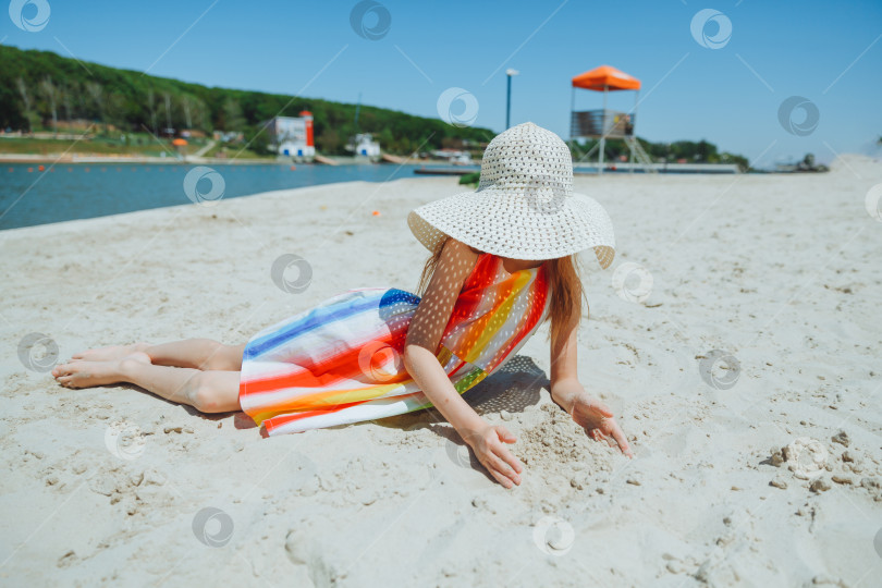 Скачать маленькая белокурая девочка в соломенной шляпке лежит на песчаном пляже. девушка загорает на песке фотосток Ozero