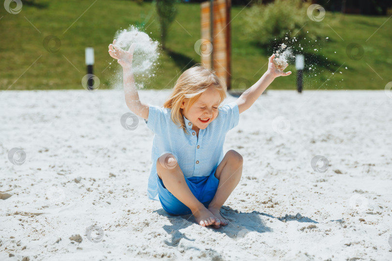 Скачать Маленький белокурый мальчик с длинными волосами сидит на песке на пляже во время летних каникул и разбрасывает песок. фотосток Ozero