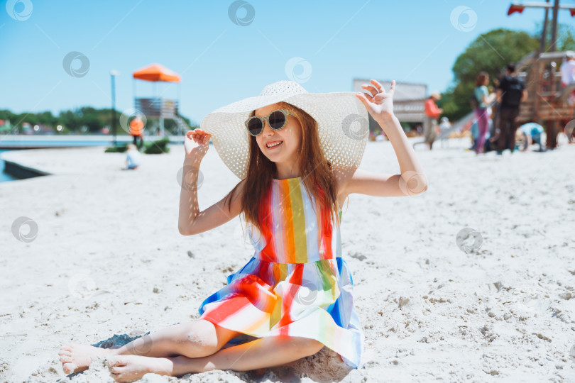 Скачать Портрет красивой маленькой девочки в полосатом платье и соломенной шляпке, сидящей на пляже фотосток Ozero