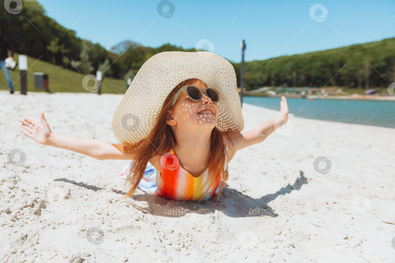 Скачать маленькая светловолосая девочка в соломенной шляпке лежит на песчаном пляже. девушка загорает на песке фотосток Ozero