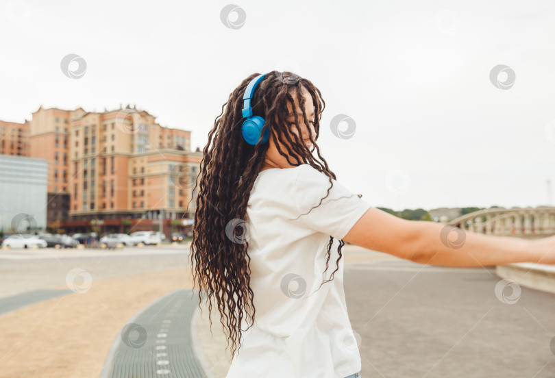Скачать Молодая жизнерадостная счастливая женщина с дредами, одетая в белую футболку, танцует, слушает музыку в наушниках, отдыхает, расслабляется в городском парке, прогуливается по аллее. Концепция городского образа жизни фотосток Ozero