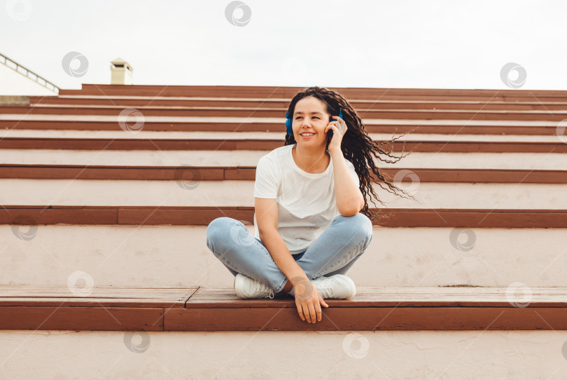 Скачать Портрет улыбающейся молодой женщины с дредами, сидящей на скамейке в парке и слушающей музыку. Прослушивание музыки в наушниках . поколение z. фотосток Ozero