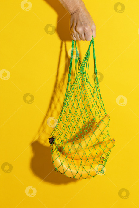 Скачать Мужская рука держит зеленую сетчатую сумку с бананами на желтом фоне фотосток Ozero