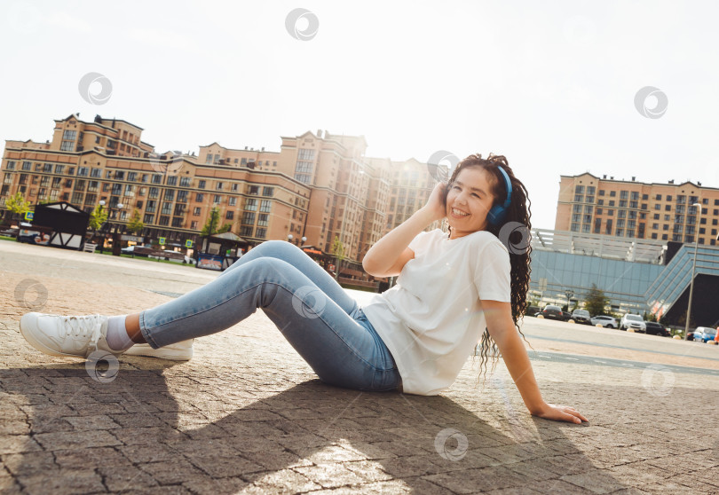 Скачать Улыбающаяся девушка с дредами в наушниках сидит на полу в парке. Счастливая молодая женщина расслабляется в наушниках, наслаждаясь музыкой. Пространство для копирования.поколение z фотосток Ozero