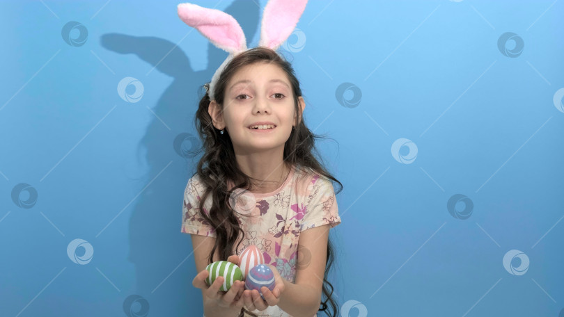 Скачать Симпатичная девушка с заячьими ушками держит в руках разноцветные пасхальные яйца на синем фоне. фотосток Ozero