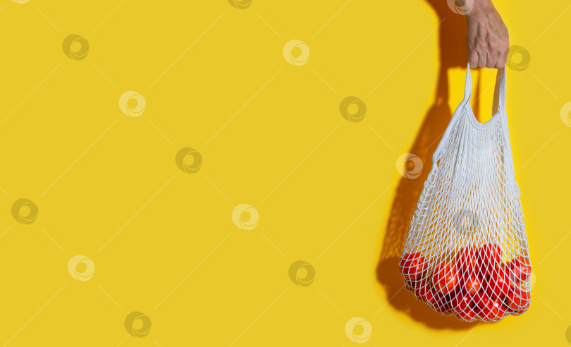Скачать Мужская рука держит белый сетчатый пакет с овощами на желтом фоне фотосток Ozero