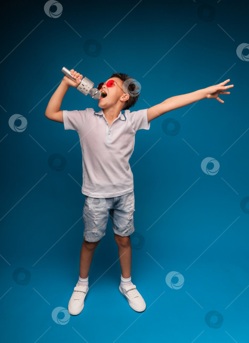Скачать мальчик поет в микрофон на синем фоне. Красивый мальчик в белой футболке и шортах стоит на синем фоне и эмоционально поет в микрофон. фотосток Ozero