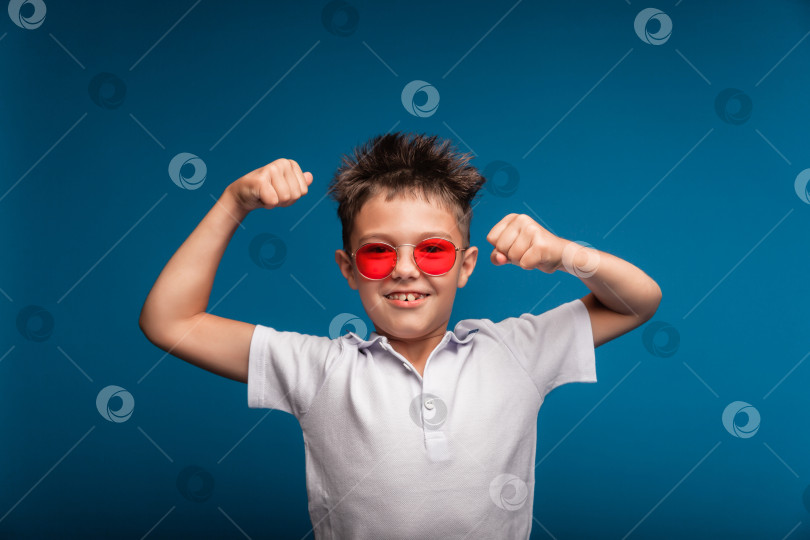 Скачать Уверенный в себе парень демонстрирует свои мускулы. Спортивный ребенок - это мальчик, демонстрирующий силу бицепсов своей руки. Школьник в очках. фотосток Ozero