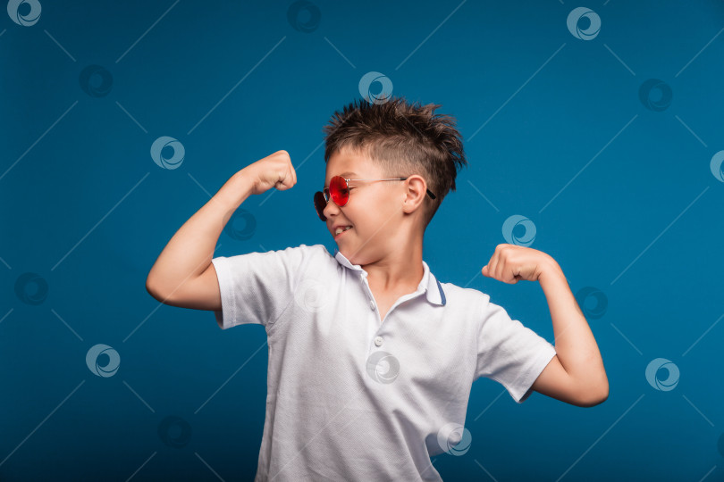 Скачать Уверенный в себе парень демонстрирует свои мускулы. Спортивный ребенок - это мальчик, демонстрирующий силу бицепсов своей руки. Школьник в очках. фотосток Ozero