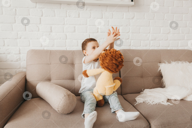 Скачать Улыбающийся мальчик в белой футболке и синих джинсах сидит на диване и обнимает мягкого игрушечного львенка фотосток Ozero