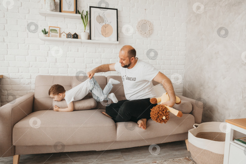 Скачать Счастливый молодой кавказский отец на диване в гостиной играет с маленьким сыном-дошкольником фотосток Ozero