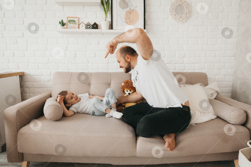 Скачать Счастливый молодой кавказский отец на диване в гостиной играет с маленьким сыном-дошкольником фотосток Ozero