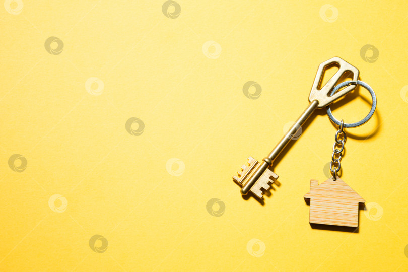 Скачать Брелок для ключей в форме деревянного домика с ключом на желтом фоне. Строительство, дизайн, проектирование, переезд в новый дом, ипотека, аренда и покупка недвижимости. Пространство для копирования фотосток Ozero
