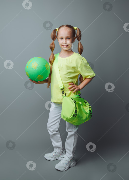 Скачать маленькая молодая девушка с зеленым мечом в руке улыбается в камеру, выделяясь на сером фоне. маленький спортсмен с мячом. детский спорт. фотосток Ozero