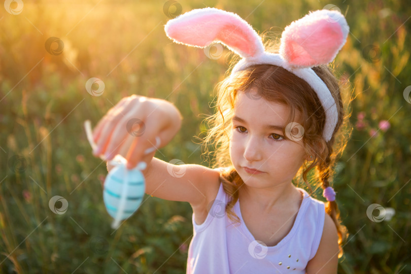 Скачать Милая забавная девочка с раскрашенными пасхальными яйцами весной на природе в поле с золотым солнечным светом и цветами. Пасхальный праздник, пасхальный кролик с ушами, разноцветные яйца в корзинке. Образ жизни фотосток Ozero