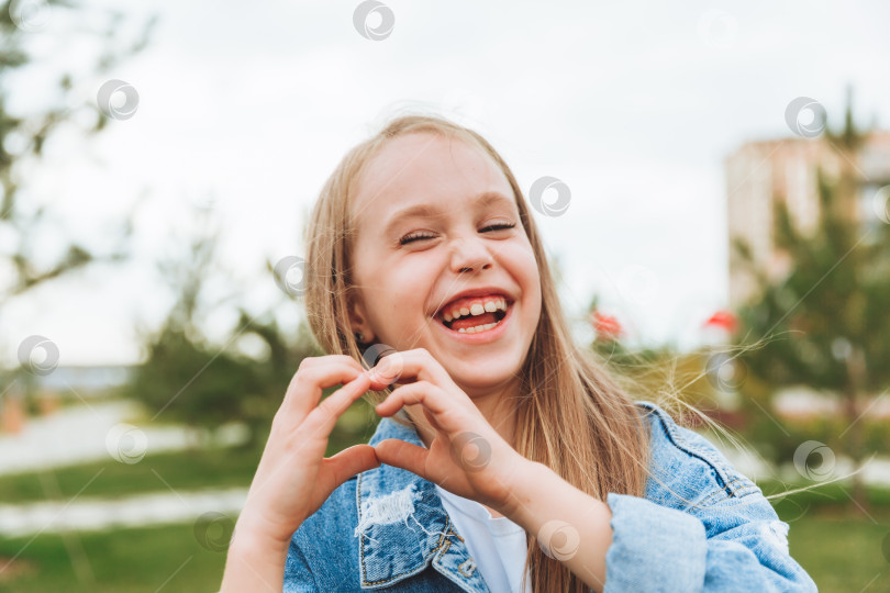 Скачать Маленькая светловолосая девочка показывает знак сердца, сделанный ее пальцами, стоя на улице в парке фотосток Ozero