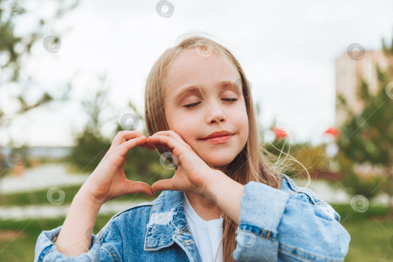 Скачать Маленькая светловолосая девочка показывает знак сердца, сделанный ее пальцами, стоя на улице в парке фотосток Ozero