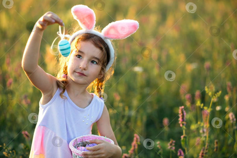 Скачать Милая забавная девочка с раскрашенными пасхальными яйцами весной на природе в поле с золотым солнечным светом и цветами. Пасхальный праздник, пасхальный кролик с ушами, разноцветные яйца в корзинке. Образ жизни фотосток Ozero
