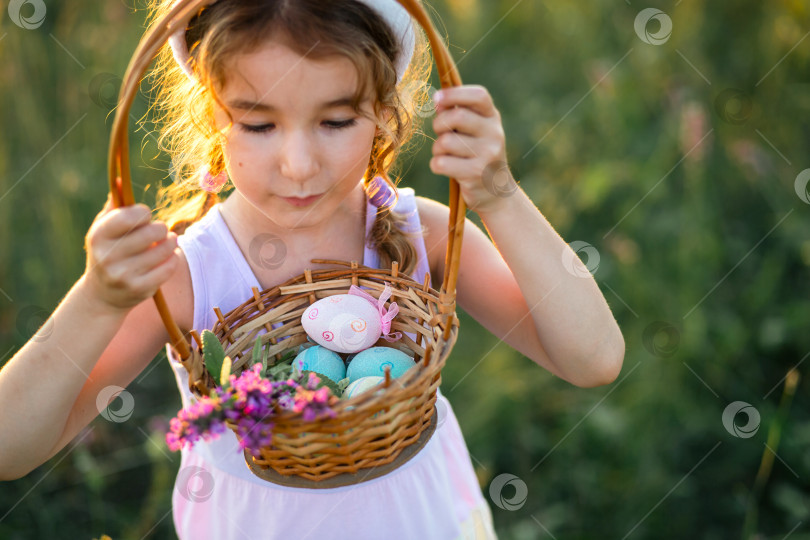 Скачать Милая забавная девочка с раскрашенными пасхальными яйцами весной на природе в поле с золотым солнечным светом и цветами. Праздник Пасхи, пасхальный кролик с ушками, разноцветные яйца в корзинке. Образ жизни фотосток Ozero