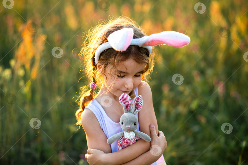 Скачать Милая 5-летняя девочка с заячьими ушками нежно обнимает игрушечного кролика на природе в цветущем поле летом, залитом золотым солнечным светом. Пасха, пасхальный кролик, детство, счастливый ребенок, весна. фотосток Ozero