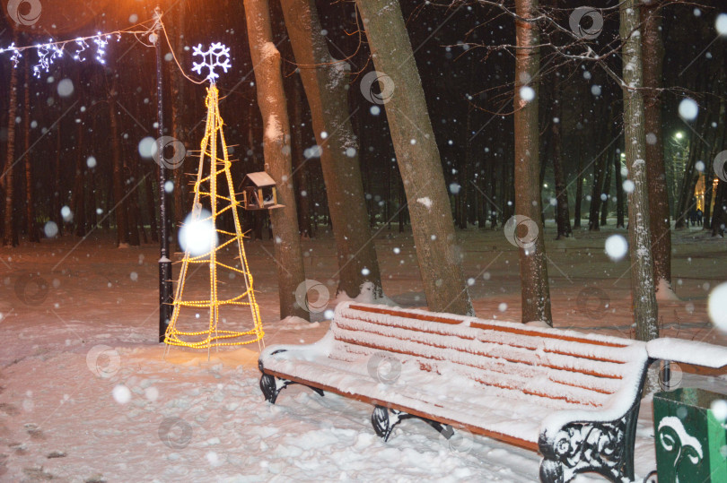 Скачать Скамейка в зимнем парке вечером, покрытая снегом, с рядом фонарей, электрической гирляндой и декоративной рождественской елкой фотосток Ozero