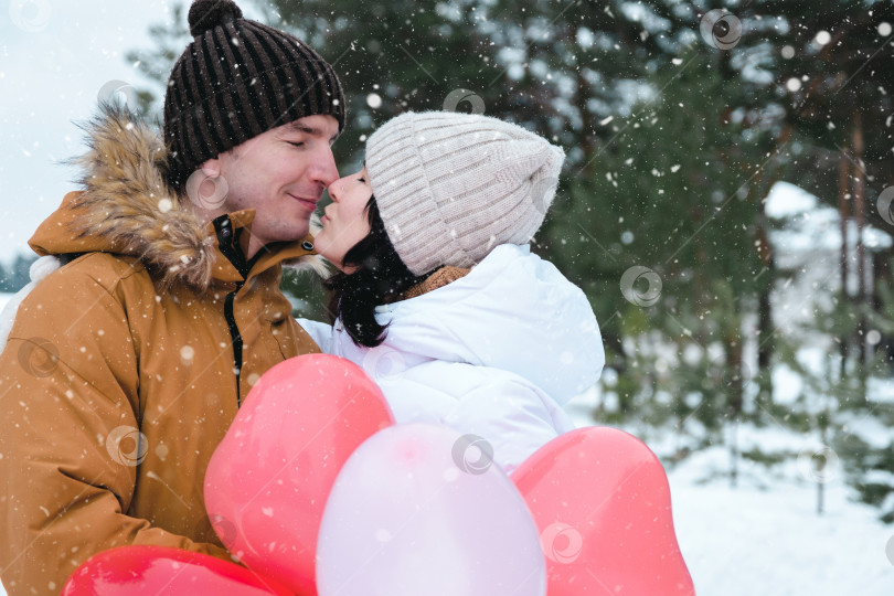 Скачать Влюбленные мужчина и женщина целуются на свидании на открытом воздухе зимой в снегу с подарком из розовых и красных воздушных шаров в форме сердца. День святого Валентина, любовь, счастливая влюбленная пара на прогулке фотосток Ozero