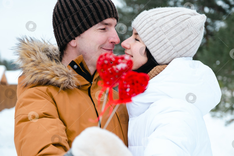 Скачать Влюбленные мужчина и женщина на свидании на открытом воздухе зимой, в снегу, с красным декором в форме сердца. День Святого Валентина, любовь, счастливая влюбленная пара на прогулке, объятия и поцелуи фотосток Ozero