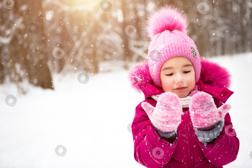 Скачать Маленькая девочка на морозе смотрит на снежные варежки и улыбается. Зима, гуляйте с малышом на свежем воздухе, снег. Теплая одежда, вязаная шапка, меховой помпон и капюшон. Игра в снежки, зимние забавы, приближается весна фотосток Ozero
