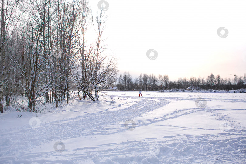 Скачать Зимний пейзаж - белый снег со следами обуви и лыж на поле. Лыжня и дорога, огибающие лес с голыми деревьями, мягкий солнечный свет. фотосток Ozero