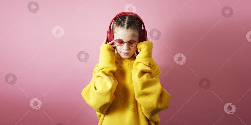 Скачать Молодая девушка в желтой толстовке с капюшоном слушает музыку в больших красных наушниках. Студийный портрет на розовом фоне. фотосток Ozero