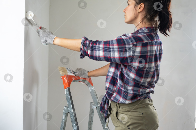Скачать Женщина с малярным валиком и белой краской раскрашивает стену дома на стремянке. Строительные работы и косметический ремонт в доме, покраска стен, тонировка, отделочные работы своими руками фотосток Ozero