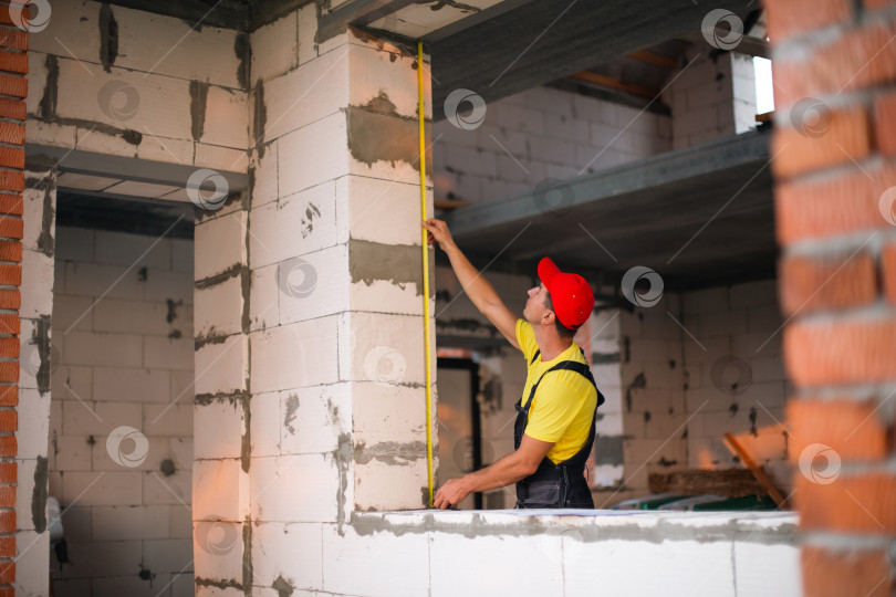 Скачать Строитель на стройплощадке измеряет рулеткой длину оконного проема и кирпичной стены. Коттедж сделан из пористых бетонных блоков, рабочая одежда - комбинезон и бейсболка фотосток Ozero