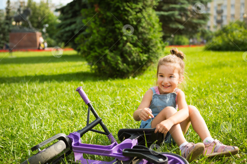 Скачать Веселая девушка сидит, отдыхая на траве в парке рядом с фиолетовым беговым велосипедом в летнее время. Детское активное развлечение, самокат для малышей, счастливый ребенок. Пространство для копирования. Летний фон фотосток Ozero