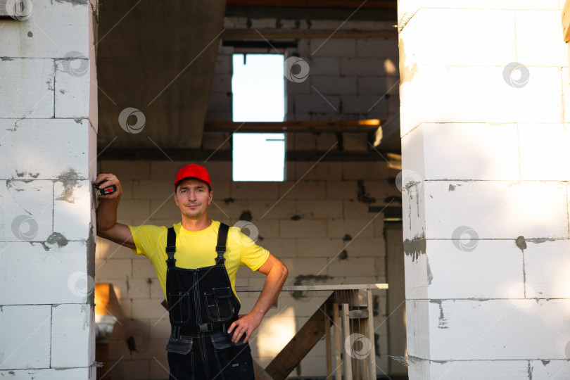 Скачать Инженер-строитель на строительной площадке измеряет рулеткой длину оконного проема и кирпичной стены. Мужчина в рабочей одежде - комбинезоне и кепке фотосток Ozero