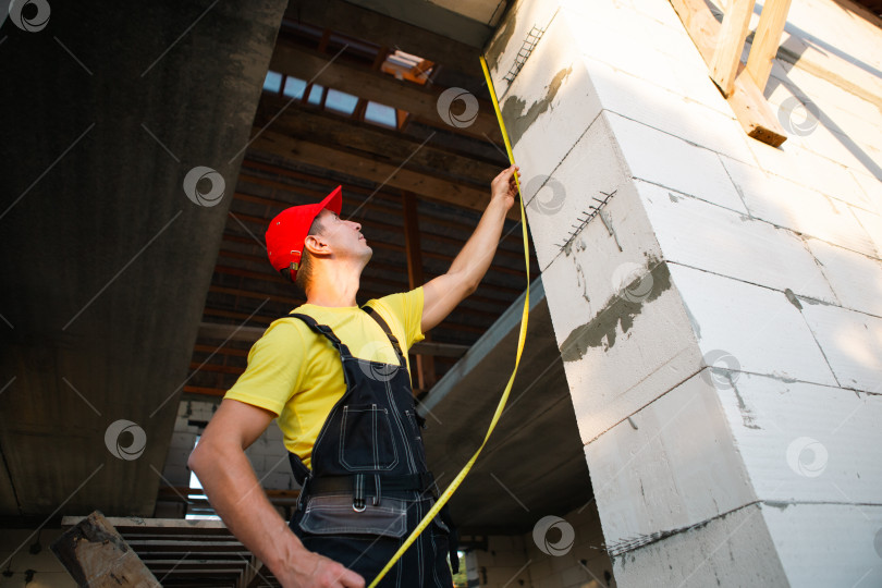 Скачать Строитель на стройплощадке измеряет рулеткой длину оконного проема и кирпичной стены. Коттедж сделан из пористых бетонных блоков, рабочая одежда - комбинезон и бейсболка фотосток Ozero
