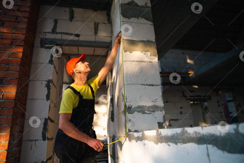 Скачать Рабочий-строитель на стройплощадке измеряет рулеткой длину оконного проема и кирпичной стены. Коттедж сделан из пористых бетонных блоков, рабочая одежда - комбинезон и бейсболка фотосток Ozero