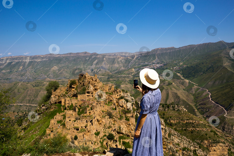 Скачать Туристка высоко в горах Дагестана любуется захватывающим панорамным видом на природу и древний заброшенный город Гамсутль. Фотографирует достопримечательности и руины, делает селфи на смартфон фотосток Ozero