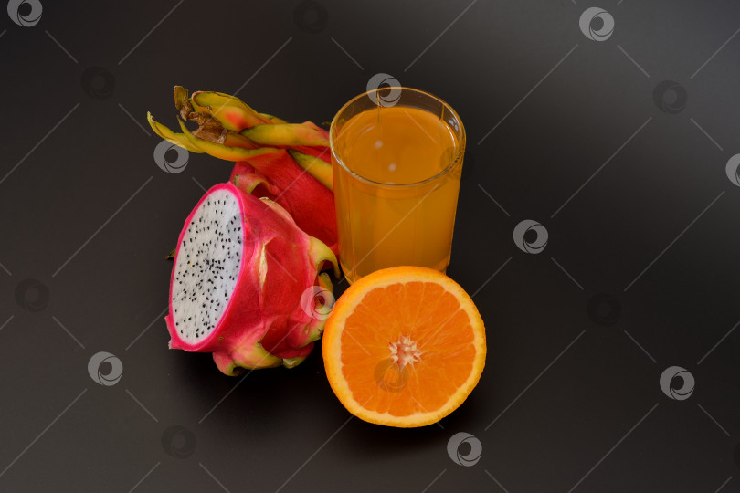 Скачать Высокий стакан фруктового сока на черном фоне, рядом со спелыми ломтиками питайи и апельсина. фотосток Ozero