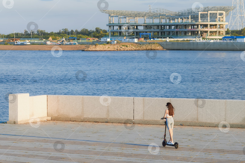Скачать A girl rides an electric scooter along the river embankment against the backdrop of the construction of an international cable car terminal. Summer evening on the Amur River фотосток Ozero