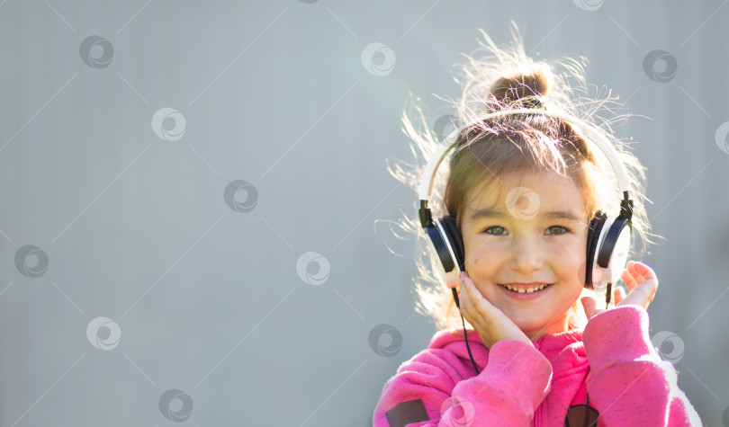 Скачать Жизнерадостная девушка в наушниках и розовой толстовке слушает музыку и смеется. Открытый, серый фон фотосток Ozero