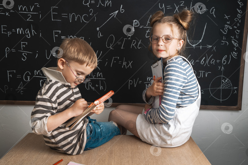 Скачать Мальчик и девочка сидят за школьной партой и изучают формулы на меловой доске. фотосток Ozero