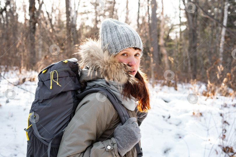 Скачать Пеший туризм: Девушка в теплой шапке с рыжими волосами и большим рюкзаком идет пешком по зимнему лесу. фотосток Ozero