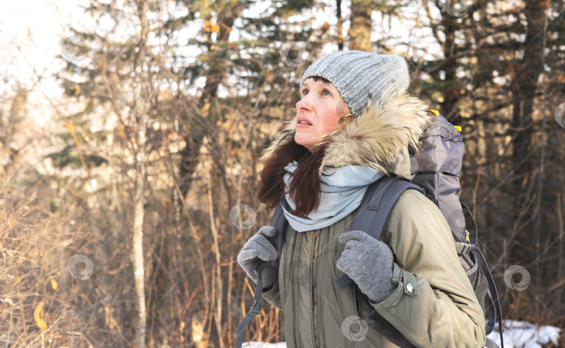 Скачать Пеший туризм: Девушка в теплой шапке с рыжими волосами и большим рюкзаком идет пешком по зимнему лесу. фотосток Ozero